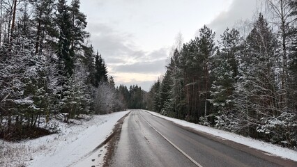 Obraz premium An asphalt road with markings, signs and sandy roadsides runs through a mixed forest. Snow is lying on the ground, roadsides and tree branches. Clear winter evening and colorful sunset sky