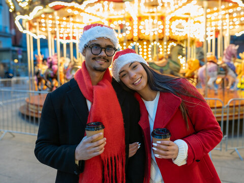 Happy young couple dressed in festive santa hats, holding coffee cups, embracing and smiling in an outdoor christmas market - Powered by Adobe