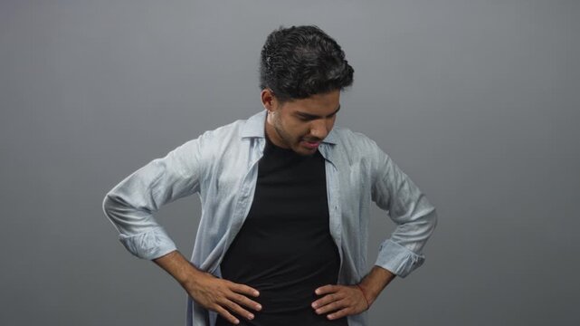 Man touching his abdomen and adjusting an open shirt while looking down in a gray studio setting; discomfort.