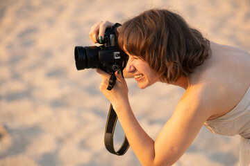 Young woman smiling and focused while holding a dslr camera, capturing golden-hour beach scenes on warm sand, backlit, relaxed and passionate about photography hobby