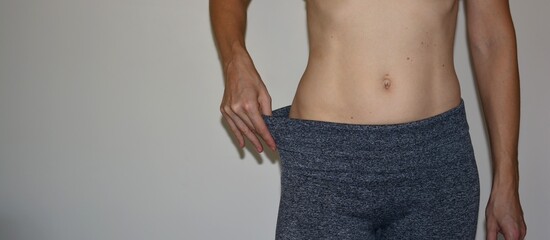 Woman showing weight loss by pulling loose sports leggings around her waist. Close-up of slim stomach and oversized pants demonstrating body changes, dieting progress and fitness results.