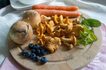 A colorful assortment of chanterelle mushrooms, blueberries, carrots, onions, and herbs arranged on a wooden board. A fresh seasonal still life for healthy eating, cooking, and organic produce concept