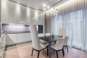 Bright and stylish kitchen features a sleek dining table surrounded by white chairs. Elegant light fixtures hang overhead, creating a warm atmosphere in the modern space.