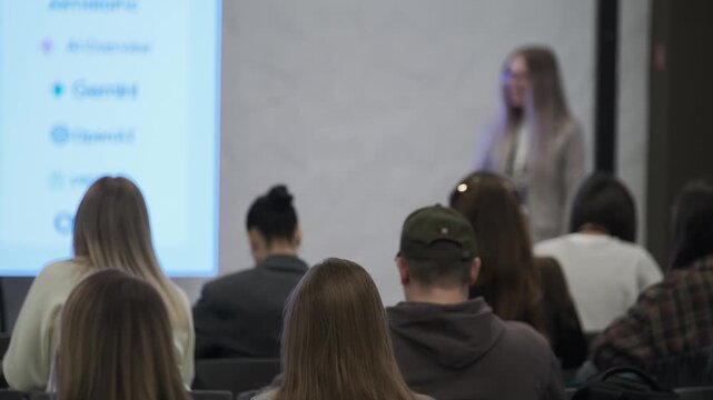 Speaker at front of room addresses audience while attendees listen, blurred projection screen creates professional event atmosphere in business presentation.