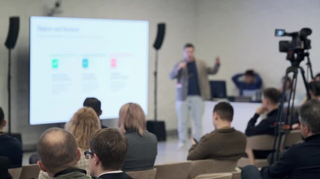 Audience watches presenter as slides glow on large screen while camera records event and microphones capture discussion