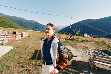 Fototapeta premium Woman in mountains village standing outdoors with backpack and jacket, sunlight on face and eyes closed enjoying nature and travel, casual style in peaceful rural landscape.