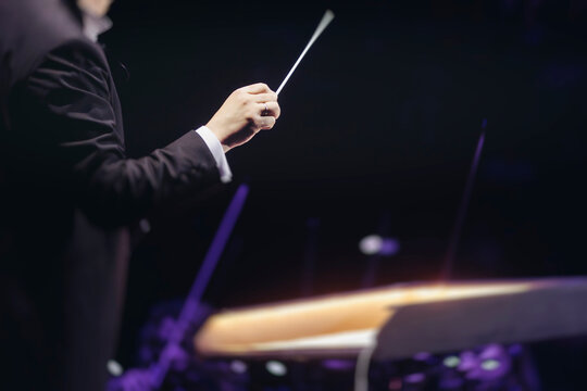 Male conductor behind the podium directing orchestra on stage with pupitre, musicians and choir, the philharmonic venue hall during concert, symphony orchestra director hands conducting waving baton