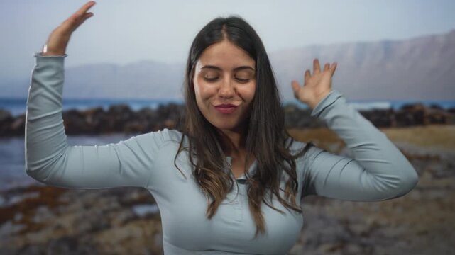 Woman with raised hands dancing on rocky seaside beach by ocean horizon under open sky; joyful spontaneity.