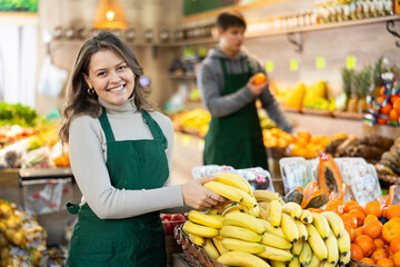 Positive young female seller holding bananas standing by counter in fruit and vegetable market