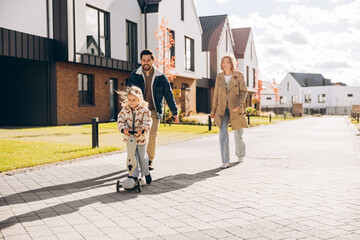Happy smiling family, woman and man walking on street with daughter riding scooter