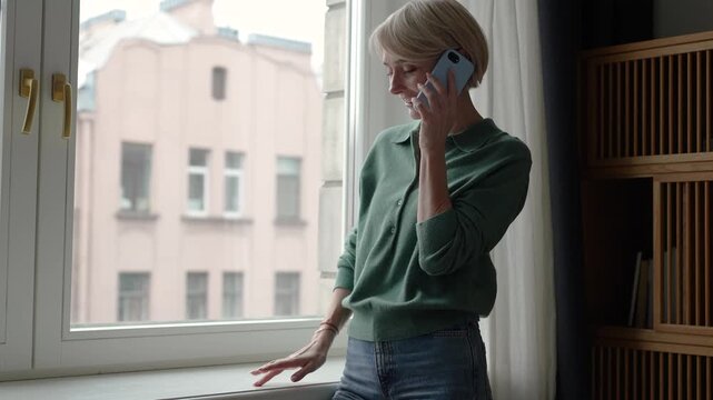 Beautiful middle aged woman having pleasant conversation on phone, standing beside window and talking with friend on smartphone. Positive female enjoying chatting on cellphone, smiling and joking.