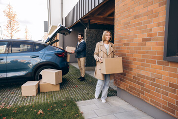 Happy couple moving into new house carrying boxes, smiling woman and man holding cardboard boxes