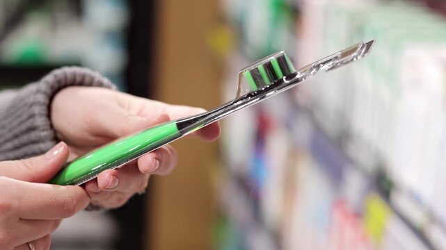 Woman holding toothbrush in a store aisle, examining the product before purchase. Close-up of hands and the toothbrush packaging with blurred shelves of oral care products in the background.