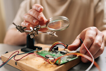 Close-up hands of technician focusing on intricate details of circuit board on wooden base with magnifying glass, using helping hands tools and wires during electronic repair or assembly process.