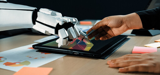 A creative workspace showing a robotic arm and human hand interacting with a touchscreen device, highlighting human-robot collaboration in design and innovation processes.Noogenesis