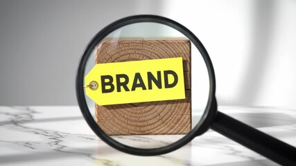 A wooden block with a yellow tag labeled BRAND is examined under a magnifying glass on a white marble surface.
