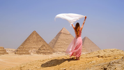 Woman enjoying her vacation in Egypt, standing joyfully in front of the pyramids with a flowing scarf in the desert breeze.