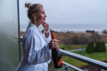 Fototapeta premium Happy Young Woman in White Bathrobe Holding Bottle of Sparkling Wine on Hotel Balcony Overlooking Beach in Parnu Estonia.