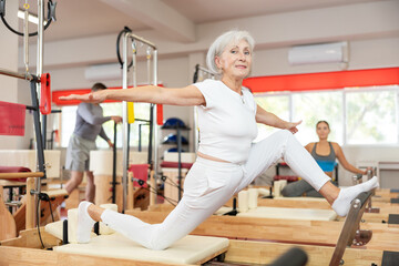Elderly woman in sportswear training stretching using pilates machine in fitness studio..