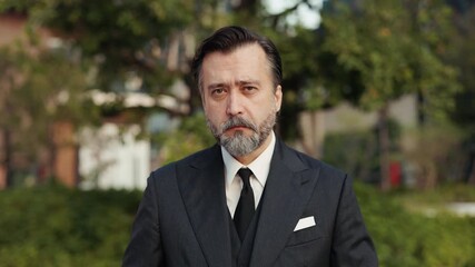Portrait of mature businessman turns head to camera in downtown park slow motion. Serious man company CEO with grey beard represents corporate success outdoor office building