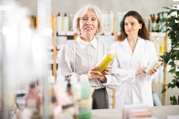 Mature woman choose shampoo, buys product in drug store. Checking for absence of GMOs, age restrictions and recommendations for use. Woman pharmacist offers product, promotes product of European brand