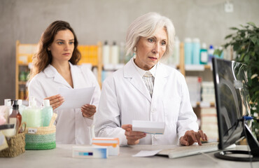 Obraz premium Elderly woman pharmacist works at a computer against the background of a female assistant. Female pharmacists take stock of pills and medicines in pharmacy
