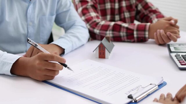 Client diligently signing important mortgage documents at the closing table next to a miniature house model and calculator symbolizing property investment