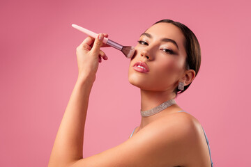 Obraz premium Gorgeous young woman applying makeup with a brush against a soft pink background, showcasing graceful beauty