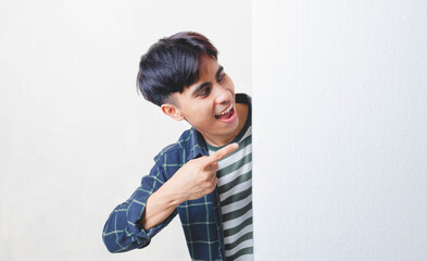 Cheerful Asian man pointing at blank copy space while peeking from behind a white wall, creating a playful promotional concept suitable for ads, marketing, and announcements.