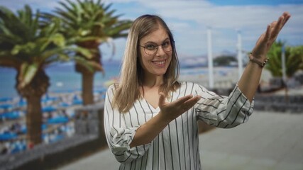 Woman wearing eyeglasses smiling while gesturing open hands on street by beach with palm trees; happiness.
