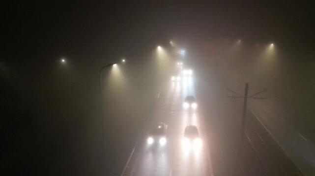 Foggy urban street scene at night with illuminated vehicles moving through mist, showcasing gradual light intensity and atmospheric depth, camera pans across the roadway