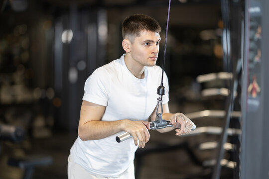 Focused young guy in white sportswear training arms on cable machine at gym, performing triceps pushdowns as part of workout routine