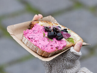Hand holding beetroot spread sandwich topped with olives and herbs at Prague Naplavka winter market, showing seasonal cold weather and cozy outdoor food atmosphere.