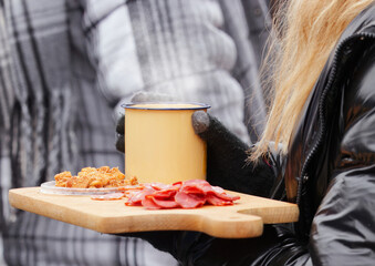 Close-up of a vendor holding a wooden board with sausage slices, cracklings, and a steaming enamel mug on a cold winter day at Naplavka farmers market.