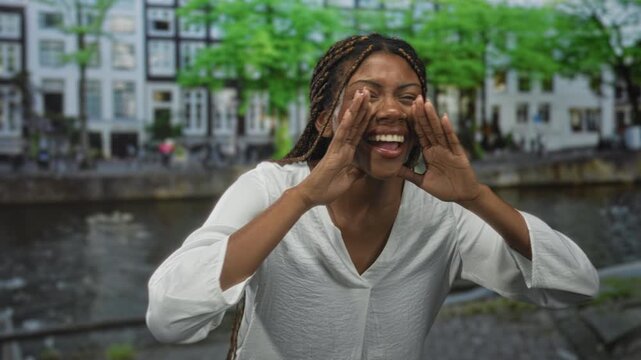 Woman face cups hands to mouth shouting on a street canal in amsterdam wearing a white blouse, braids visible; playful joy.