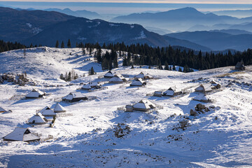 View of the scattered, traditional wooden shepherd huts of the Velika Planina settlement nestled in a snow-covered alpine pasture, with layers of blue, hazy mountain ranges in the background