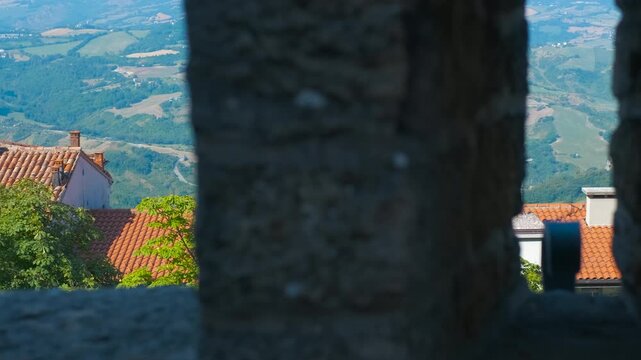 Observing rolling hills and rooftops through castle battlements. Stone battlements frame the view of a picturesque valley, featuring red-tiled rooftops nestled among green hills scene