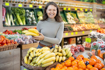 Young woman shopper in casual clothes chooses bananas in vegetable shop