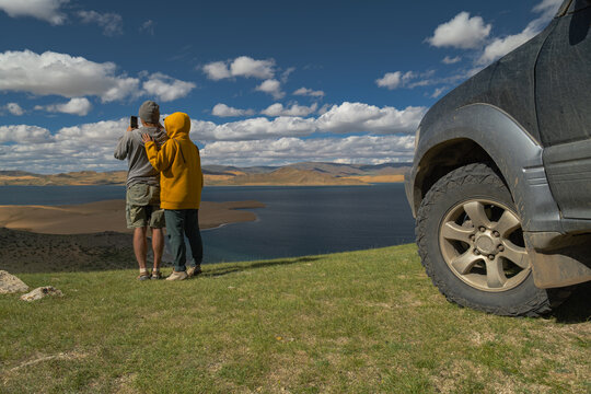 Travelers in bright clothing photograph a desert lake where sand dunes meet water. Their car, with a wheel in mid-shot, sits nearby. Backdrop of vast mountains, sandy hills, and the lake under a clear - Powered by Adobe