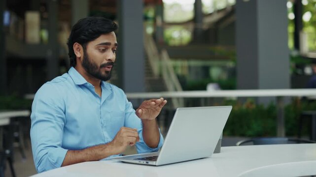 Portrait of positive Indian businessman talks with sign language on video call in street cafe. Smiling man gestures as describes situation to laptop camera in urban district