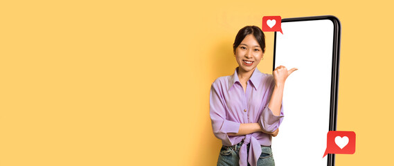 A woman stands against a bright yellow background, smiling and pointing towards a large empty smartphone screen beside her. The display features social media icons to indicate connection.