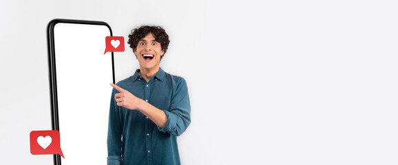 A cheerful young man stands beside a large smartphone display, pointing and smiling. He shows enthusiasm for social media engagement with heart icons appearing nearby.