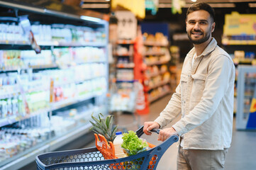 Millennial man smiling, pushing shopping cart in supermarket