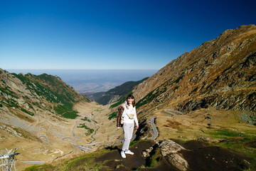 Smiling traveler woman standing on cliff edge with panoramic mountain landscape of Transfagarasan Highway, Romania. Travel concept.