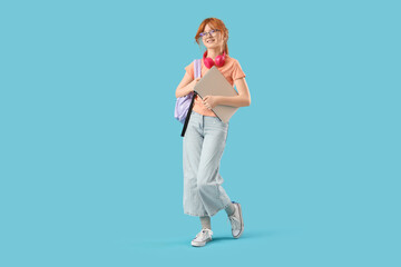 Cute schoolgirl with modern laptop, backpack and headphones walking  on blue background