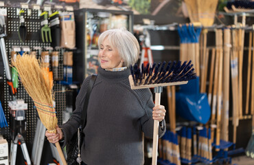 Senior woman in hardware store examines broom and besom. Client chooses and buys tool for sweeping...