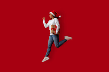 A woman wearing a Santa hat is excitedly running while holding a wrapped gift against a bright red...