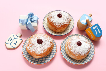 Plates with donuts, gift and dreidels for Hanukkah celebration on pink background