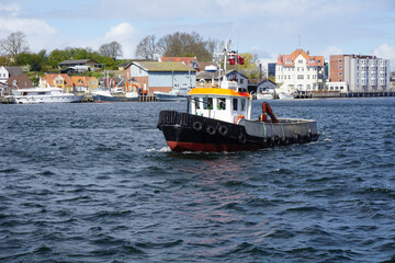 Fototapeta premium Small tugboat navigating dark blue choppy waters in Sønderborg Harbor, Denmark, with colorful waterfront buildings in the background