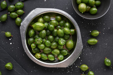 Metal plate and bowl with fresh gooseberries on black background